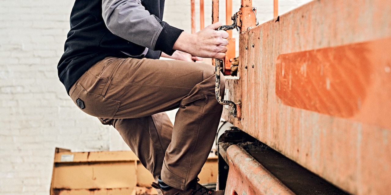 Photograph of a man wearing work cloths and climbing to the top of a forklift.