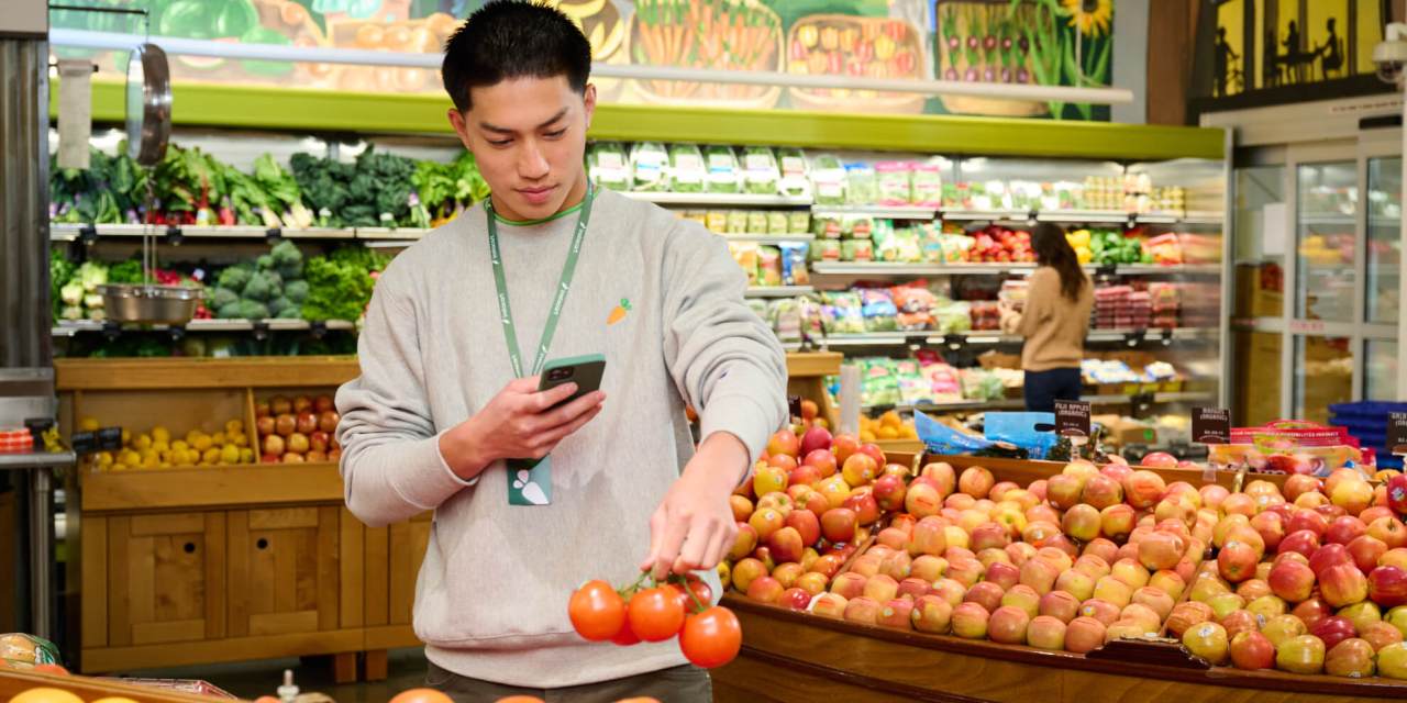 Photograph of a man picking up produce at a store and taking a picture of it with his phone.