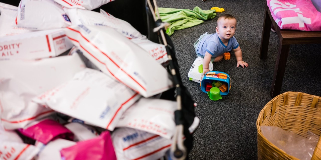 A baby crawling on the floor next to a bucket of mailers