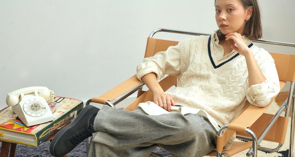 Model in a white sweater and gray pants sits in a tan lounge chair showing off black Atoms sneakers.