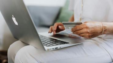 A photo of someone purchasing something on their computer. We see a person with a laptop on their lap and a credit card in their hand. Representing the purchasing of goods and the topic of the article - how retailers are reducing returns