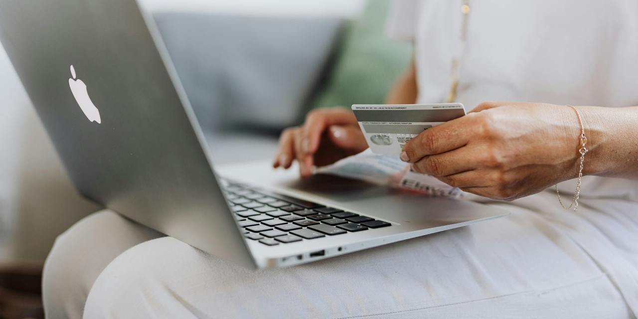 A photo of someone purchasing something on their computer. We see a person with a laptop on their lap and a credit card in their hand. Representing the purchasing of goods and the topic of the article - how retailers are reducing returns